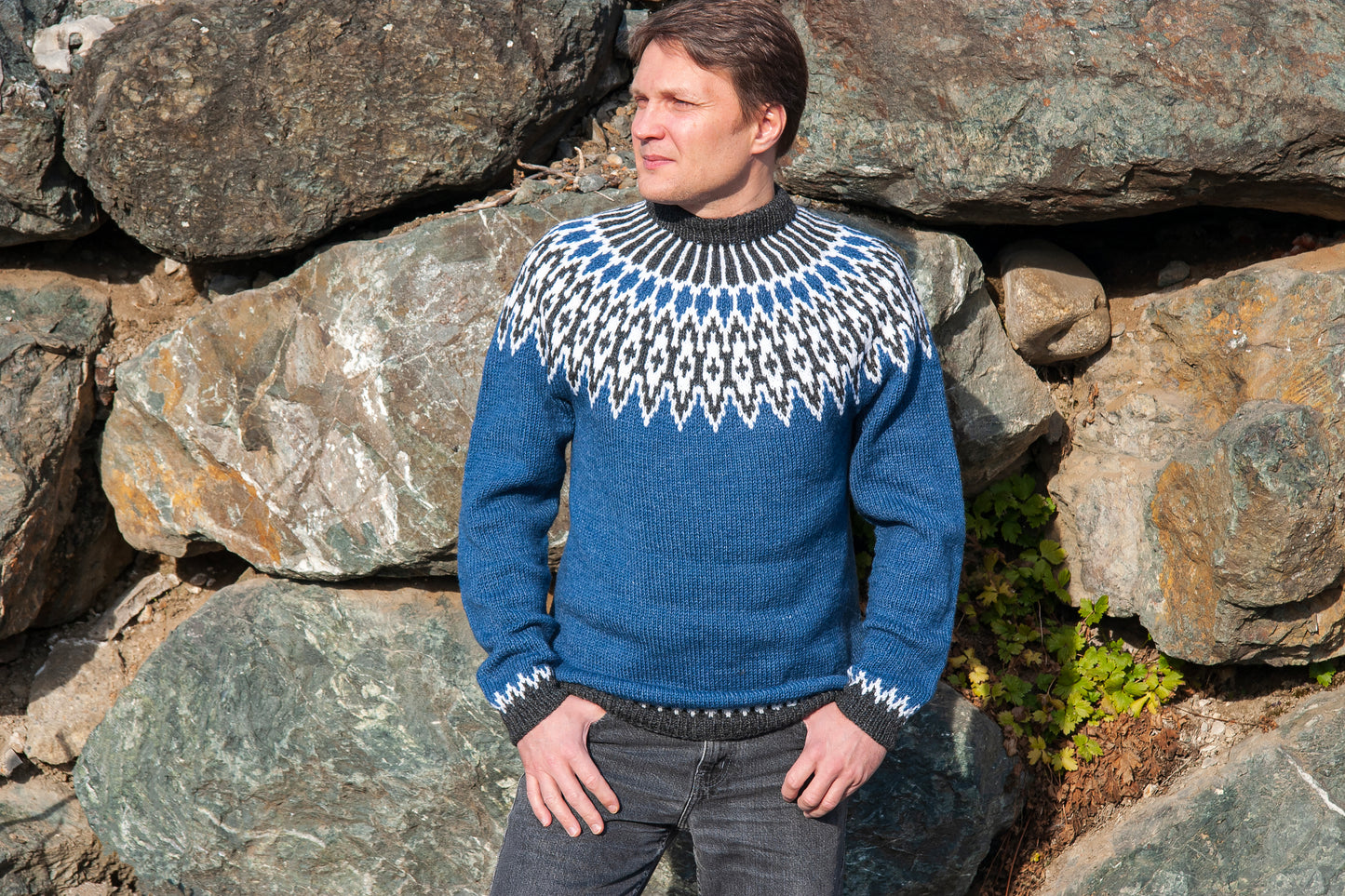 A man wearing a blue, white, and gray Odin icelandic lopapeysa sweater with a classic yoke, standing outdoors in front of large rocks.