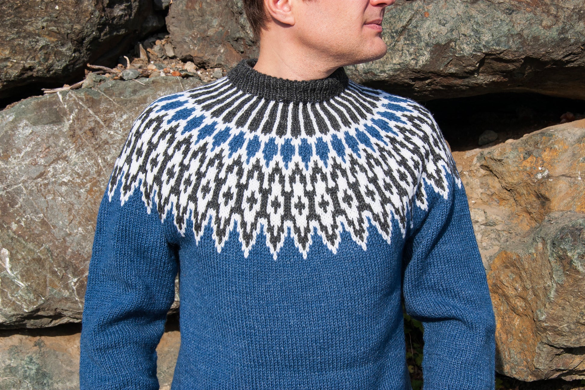 A man wearing a blue knit Icelandic lopapeysa sweater in Odin design with a black and white geometric yoke pattern, standing outdoors in front of large rocks.