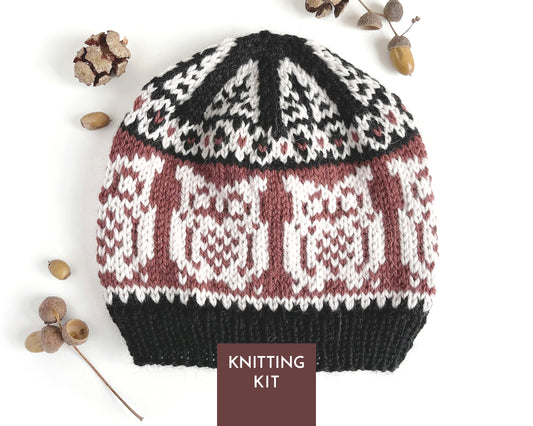 A knitted beanie hat with a Fair Isle owl pattern in black, chalk, and bordeaux, styled with acorns on a white background. A brown label at the bottom reads 'KNITTING KIT'.