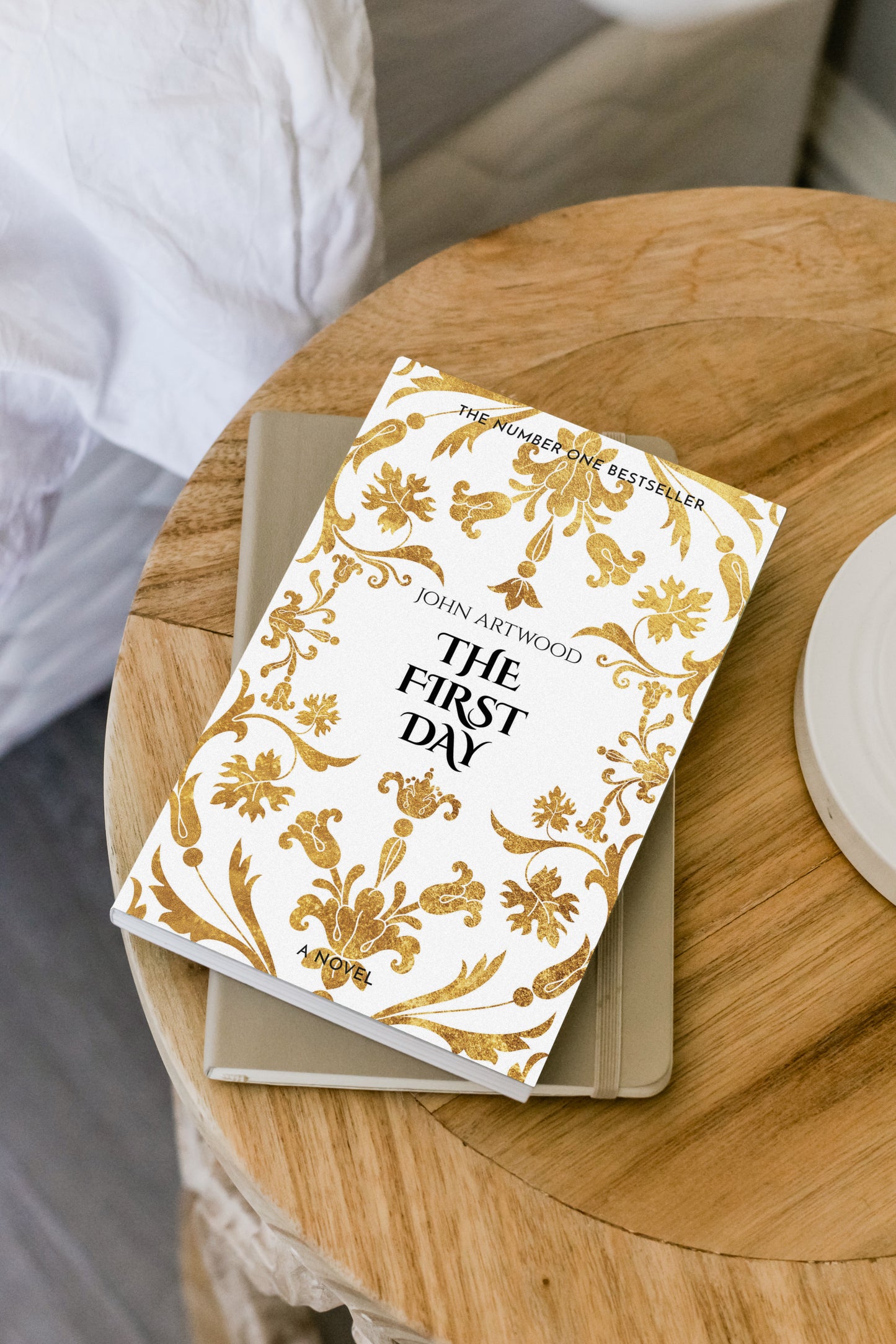 A paperback book cover featuring gold baroque ornaments on a white background, displayed on a round wooden table near a bed