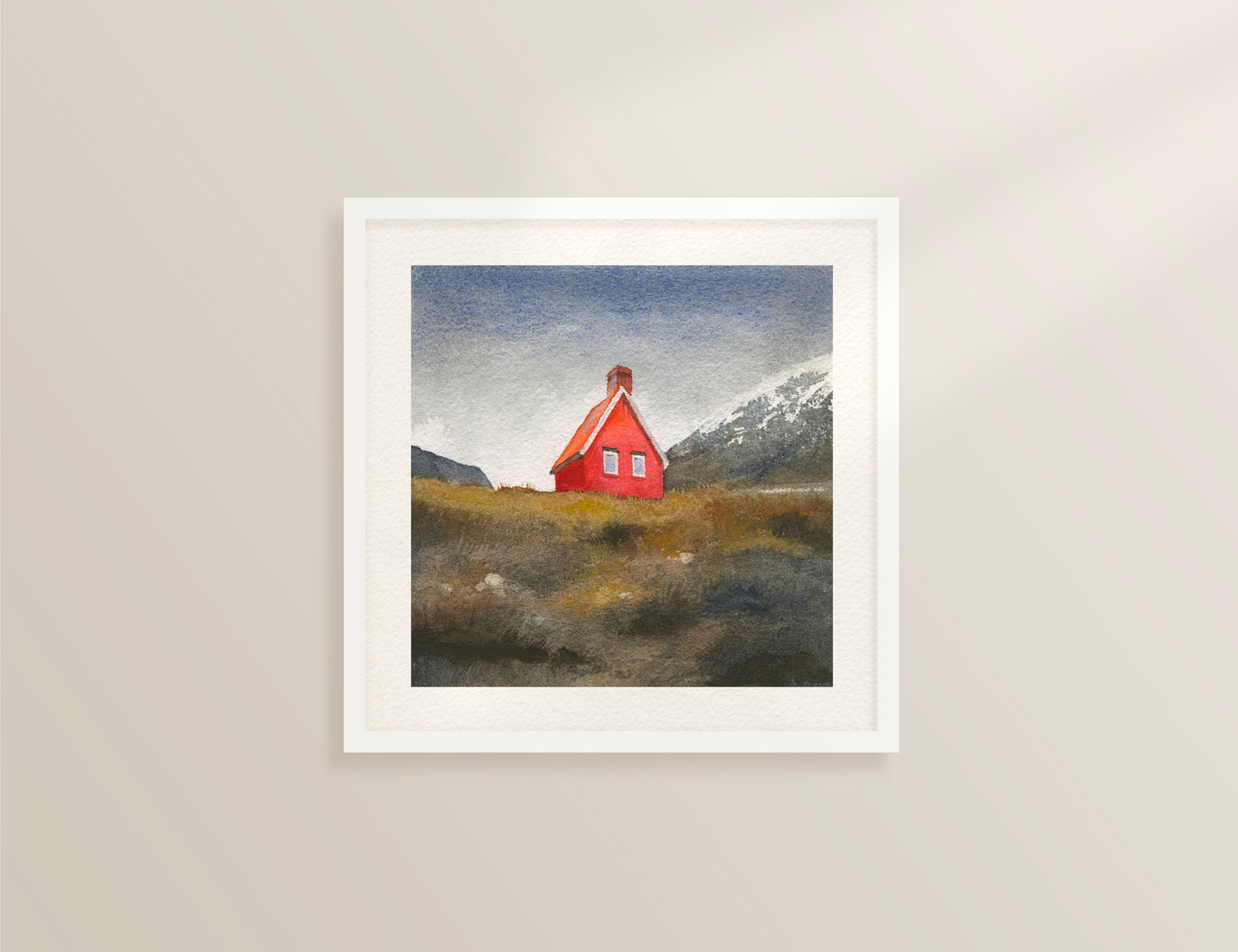 Framed watercolor painting featuring Red Cabin near Egilsstaðir, known locally as the Red Cabin of Fagridalur, stands along the road through East Iceland, framed by golden grass and distant snow-capped peaks, dispayed on a beige wall.
