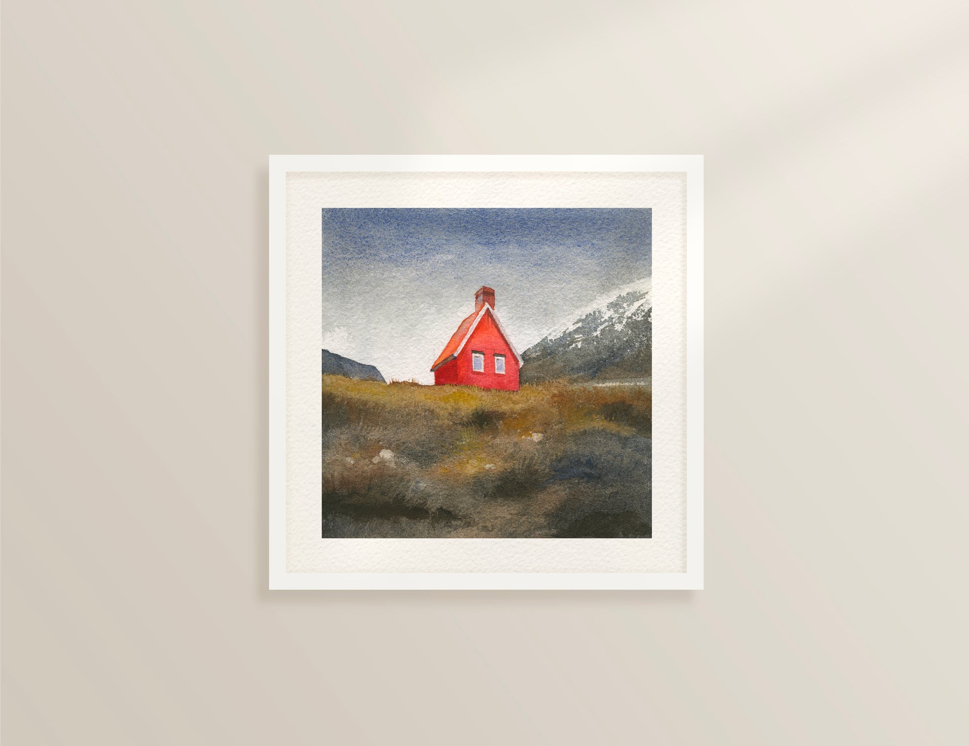 Framed watercolor painting featuring Red Cabin near Egilsstaðir, known locally as the Red Cabin of Fagridalur, stands along the road through East Iceland, framed by golden grass and distant snow-capped peaks, dispayed on a beige wall.