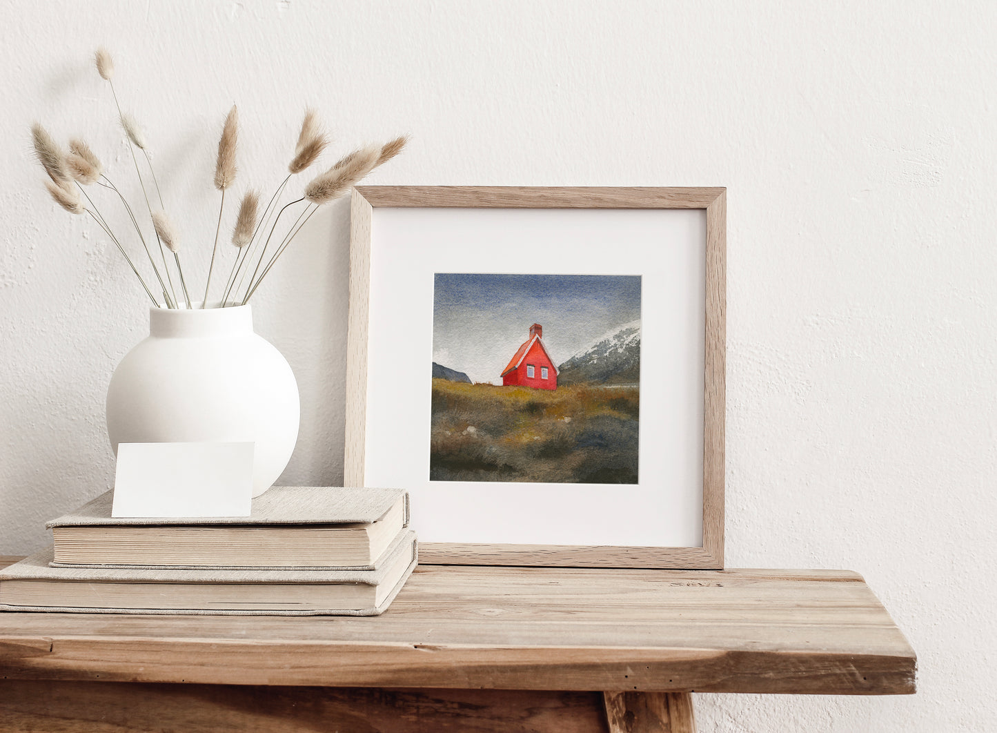 Framed watercolor painting featuring Red Cabin near Egilsstaðir, known locally as the Red Cabin of Fagridalur, stands along the road through East Iceland, displayed on a wooden table with a white vase and books.