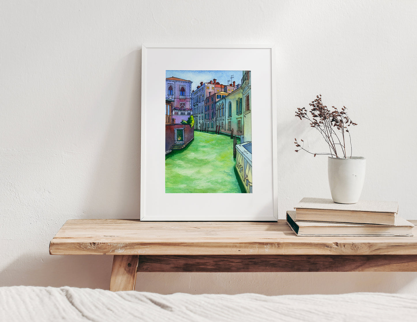 Framed watercolor artwork of of a Rio di San Polo in Venice scene on a wooden shelf with books and a plant.