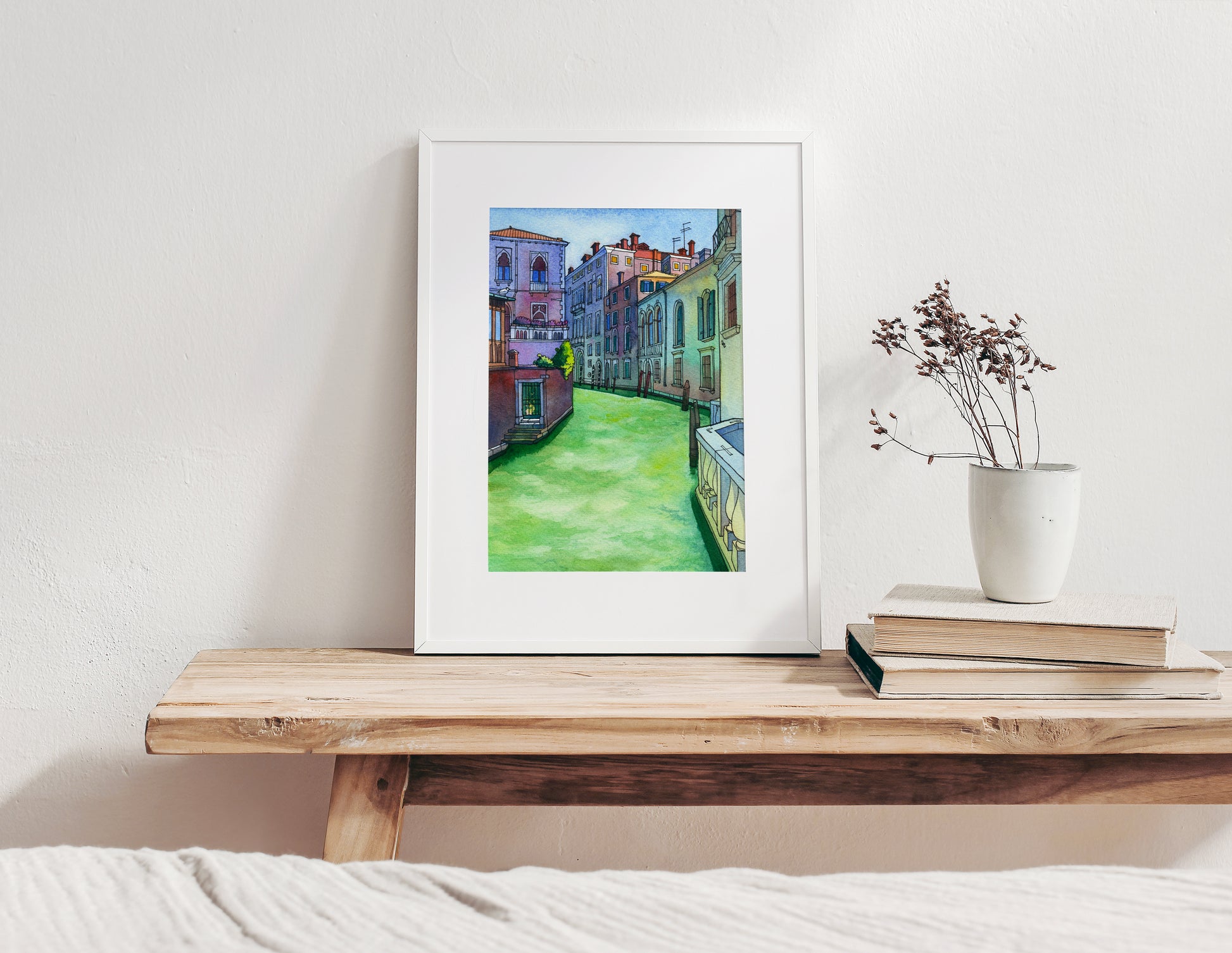 Framed watercolor artwork of of a Rio di San Polo in Venice scene on a wooden shelf with books and a plant.