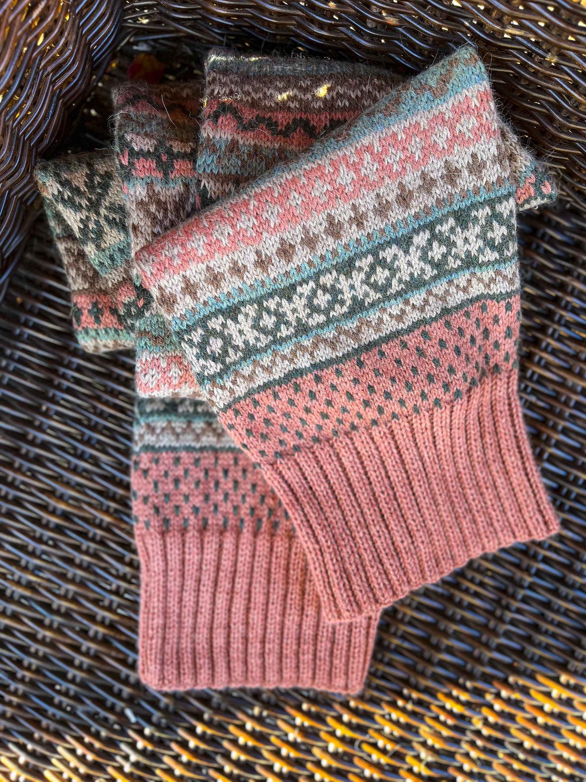 Close-up of a knitted Fair Isle scarf in dusty pink, teal, brown, and cream, arranged on a dark brown woven wicker surface.