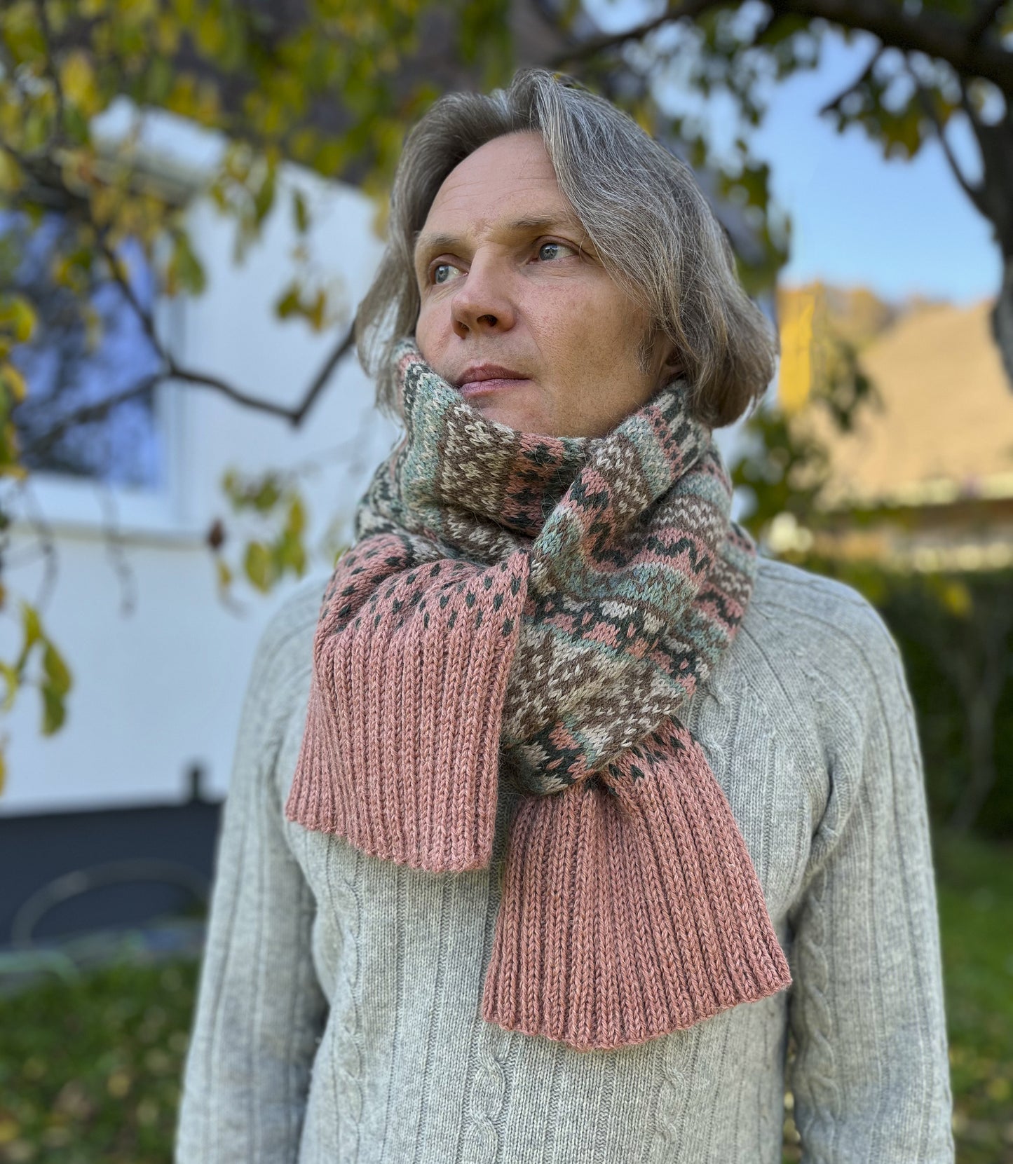 A man outdoors wearing a Fair Isle knit scarf with a multicolor pattern of green, brown, and pink, paired with a light gray cable-knit sweater.