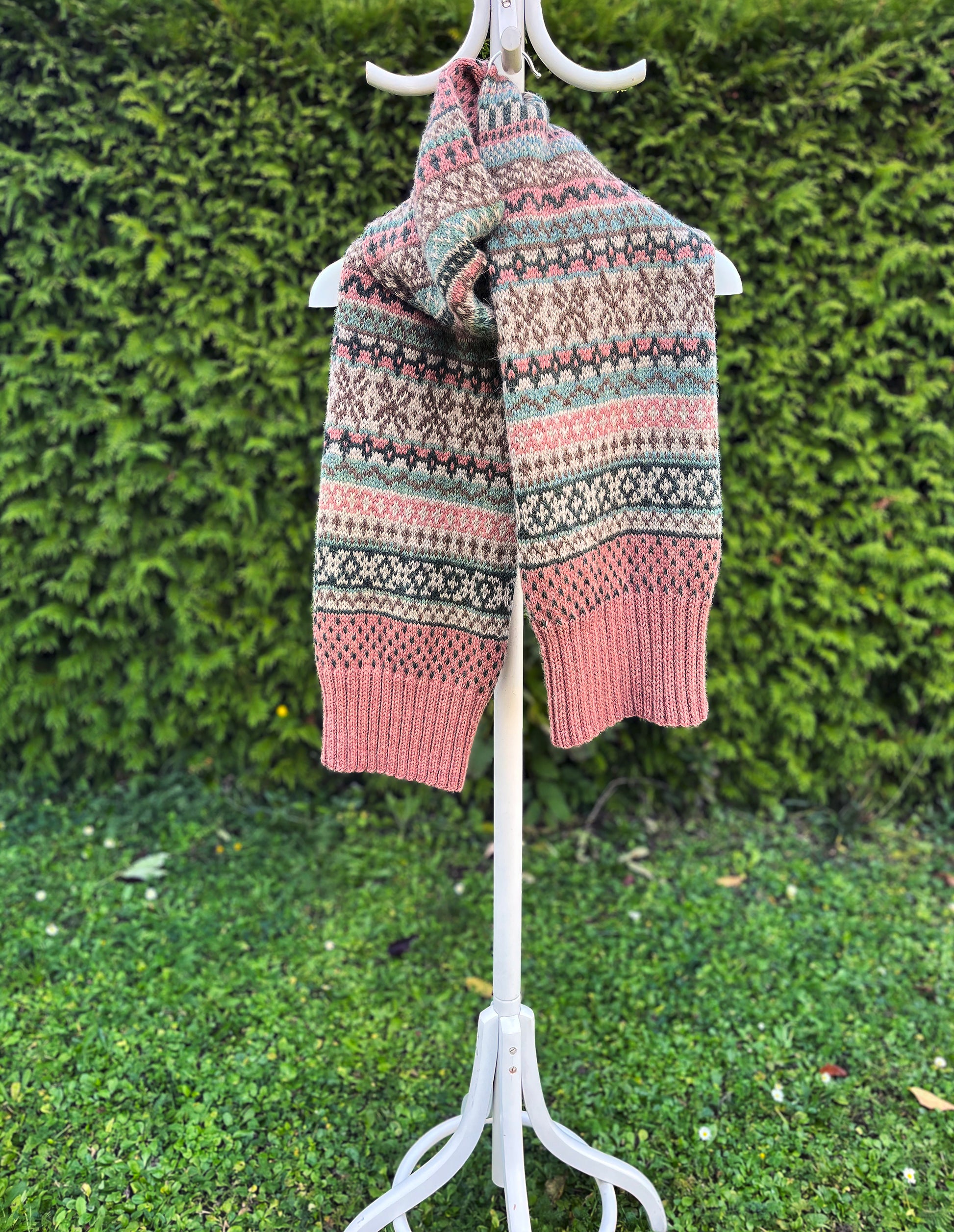 A knitted scarf with a Fair Isle pattern in dusty pink, sage green, and dark brown, hanging on a white coat rack against a green hedge background.