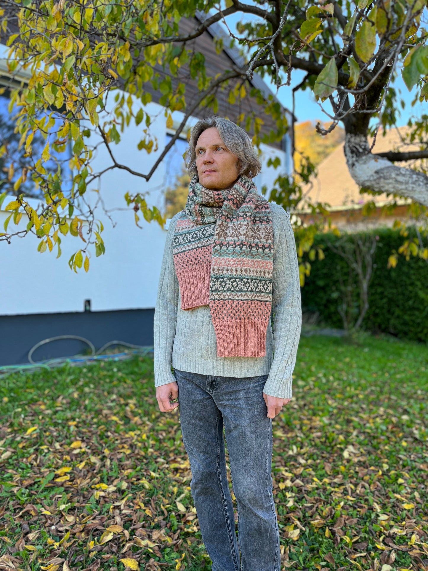 A person standing outdoors in a grassy yard under tree branches, wearing a light gray knit sweater and a multicolored Fair Isle patterned scarf with pink, green, and cream sections.