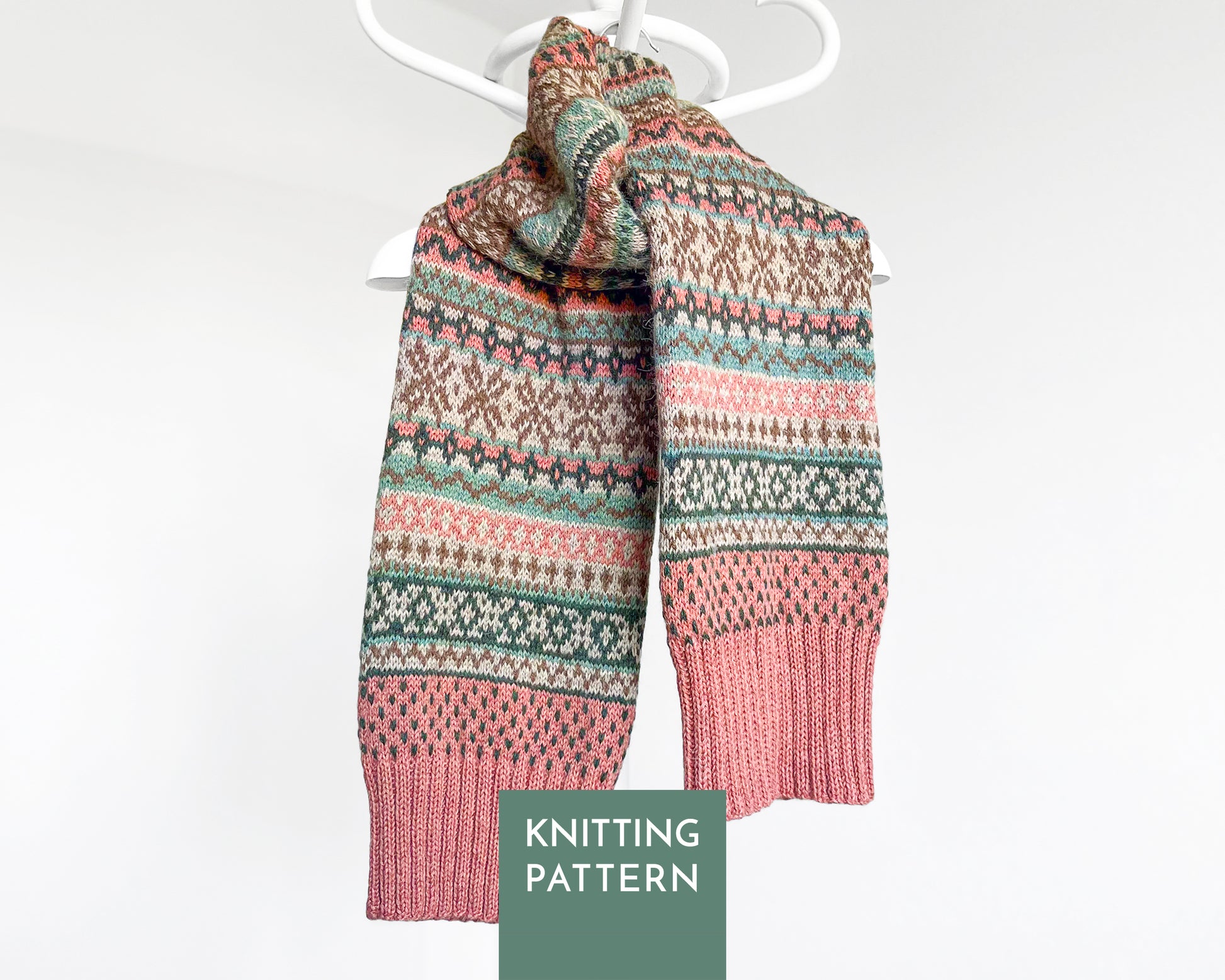 A knitted scarf with a Fair Isle pattern in dusty pink, sage green, and cream, hanging on a white hanger against a light gray background. A green label at the bottom reads 'KNITTING PATTERN'.