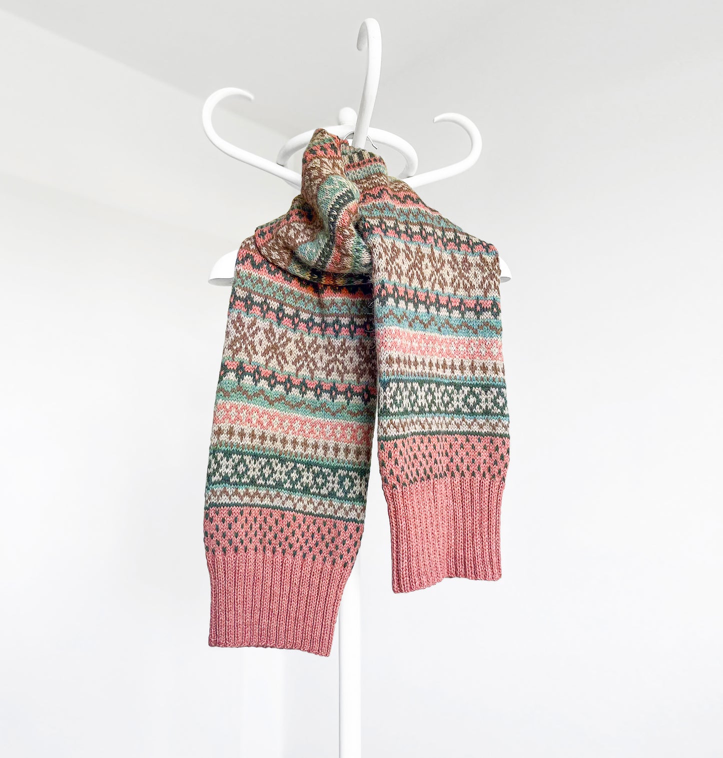A knitted scarf with a multicolor Fair Isle pattern in pink, teal, brown, and cream, draped over a white plastic hanger against a plain white wall.