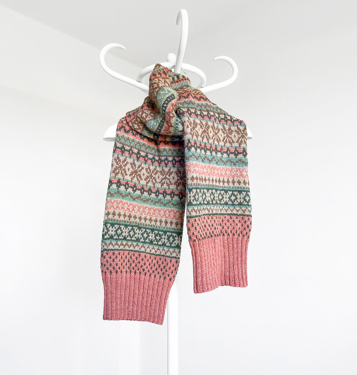 A knitted scarf with a multicolor Fair Isle pattern in pink, teal, brown, and cream, draped over a white plastic hanger against a plain white wall.