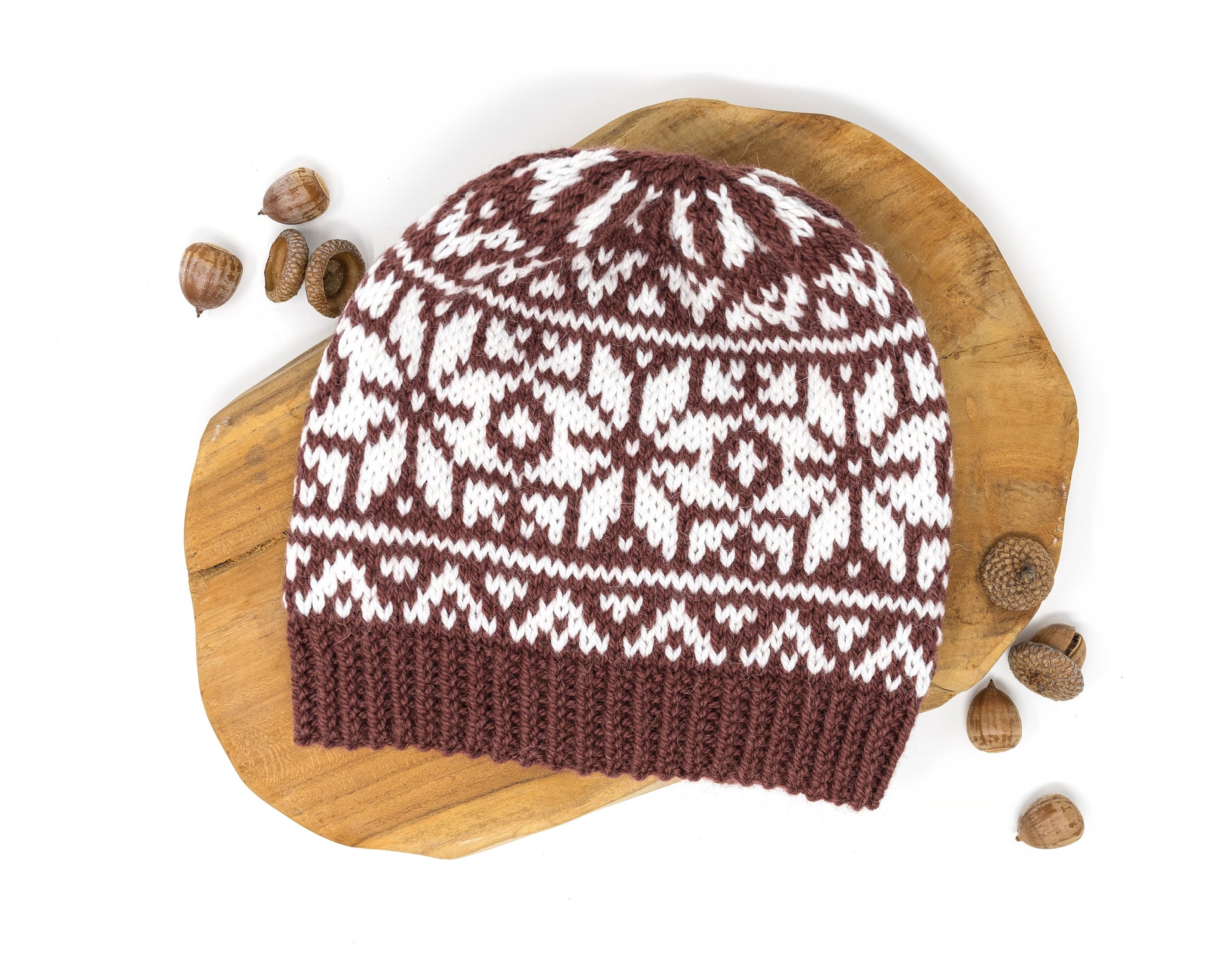 A brown and white knitted beanie hat with a classic Norwegian selburose pattern, displayed on a rustic wooden slice with scattered acorns on a white background.