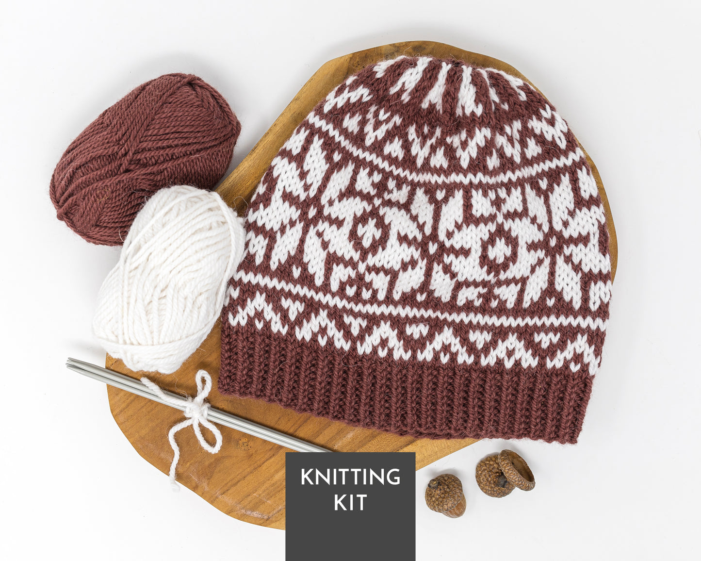 A knitted beanie with a brown and white selburose pattern is displayed on a wooden board with a metal knitting needles, two balls of yarn (one brown, one white), with a 'KNITTING KIT' label overlay.
