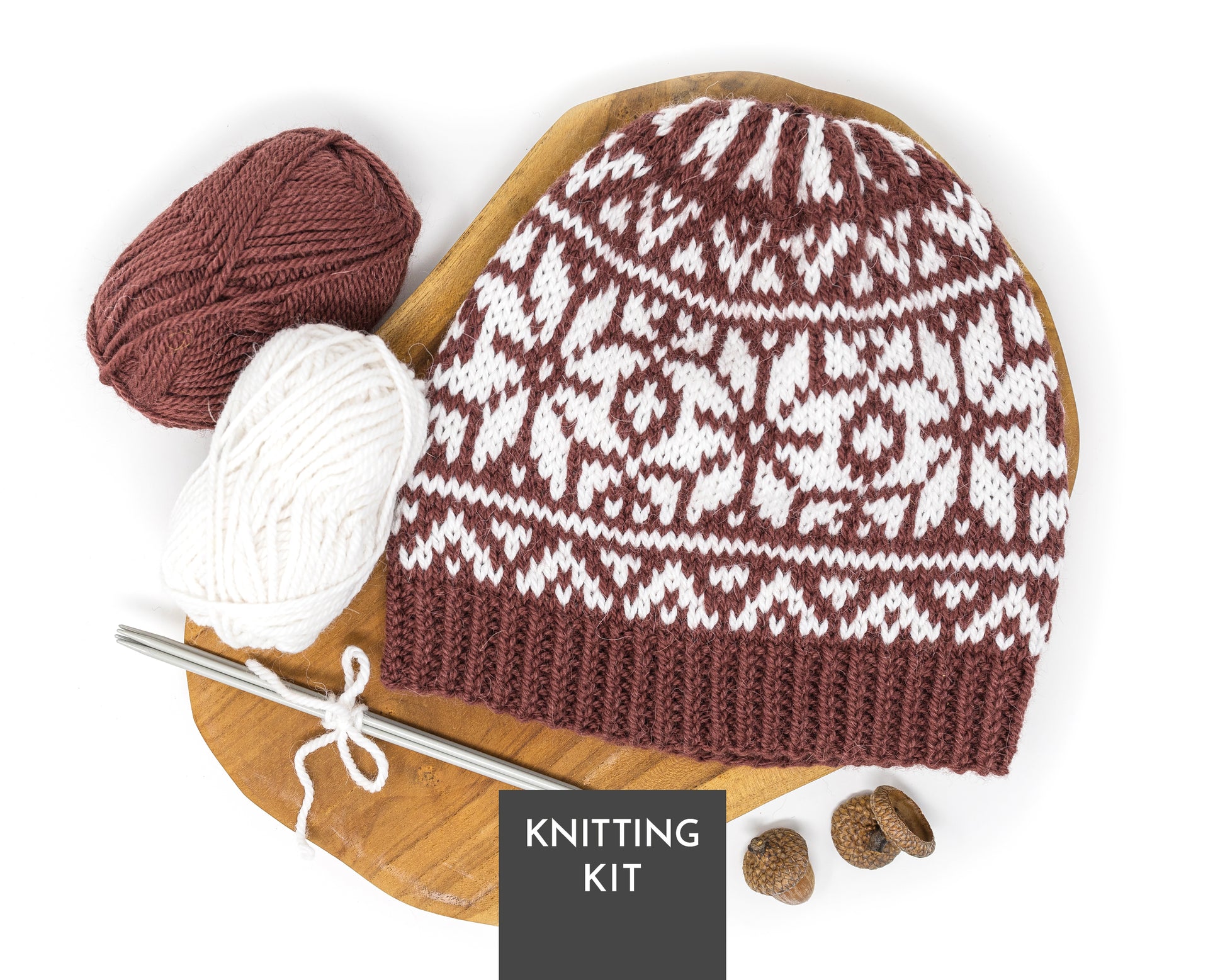 A knitted beanie with a brown and white fair isle pattern, displayed on a wooden board with a crochet hook, yarn balls in brown and white, acorns, and a 'KNITTING KIT' label on a white background.