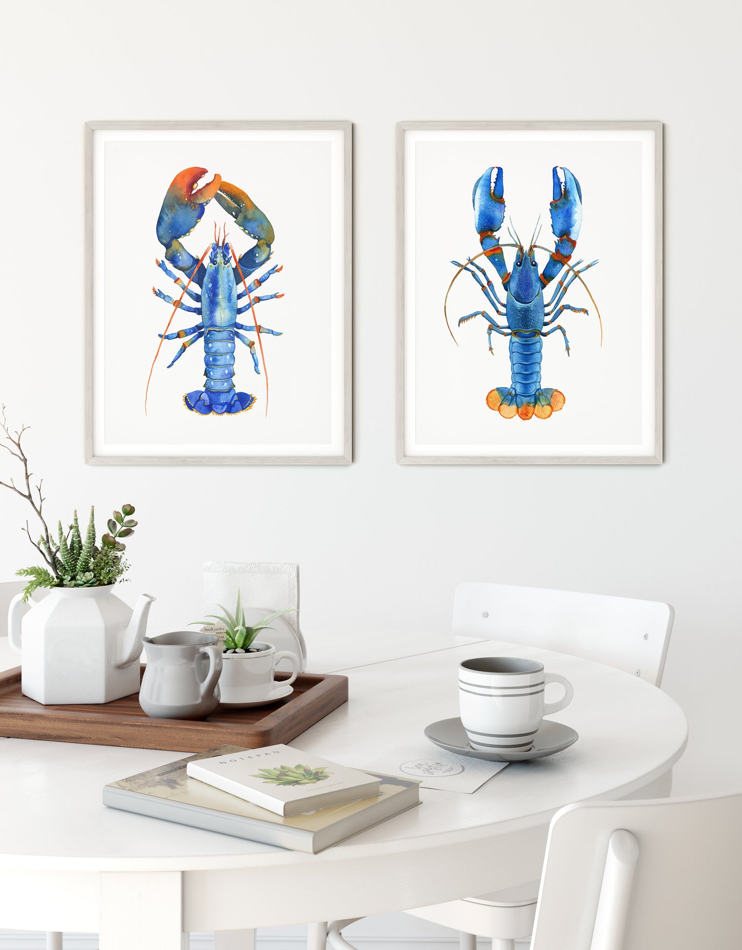 Set of two framed watercolor lobster prints hanging side-by-side on a kitchen white wall.