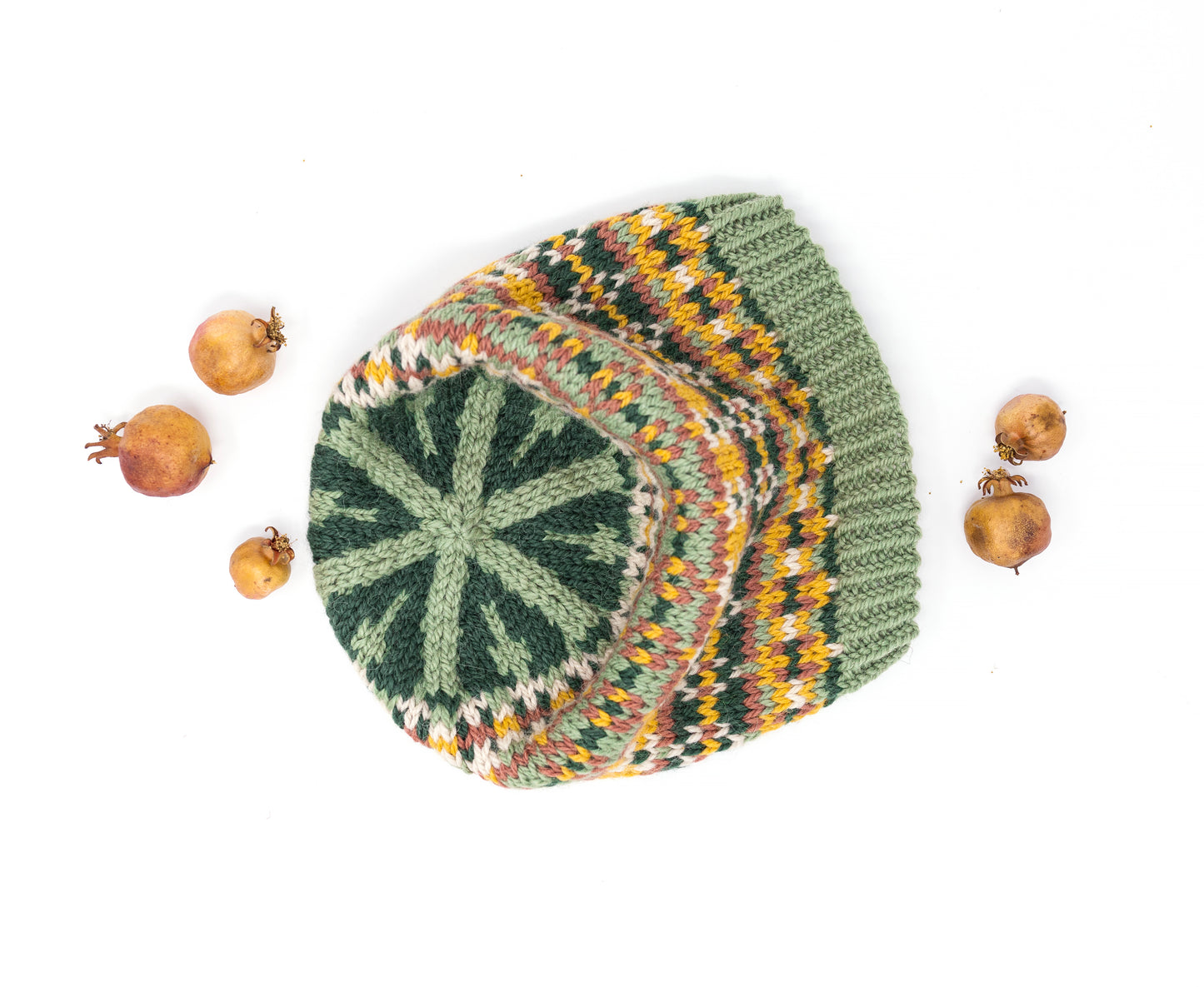 A crown of a hand-knitted beanie hat with a Fair Isle style pattern in sage green, dark green, yellow, orange, and beige, styled flat-lay on a white background with dried pomegranates.