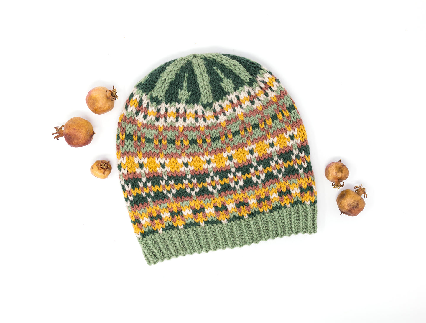 A hand-knitted beanie hat with a Fair Isle style pattern in sage green, dark green, yellow, orange, and beige, styled flat-lay on a white background with dried pomegranates.