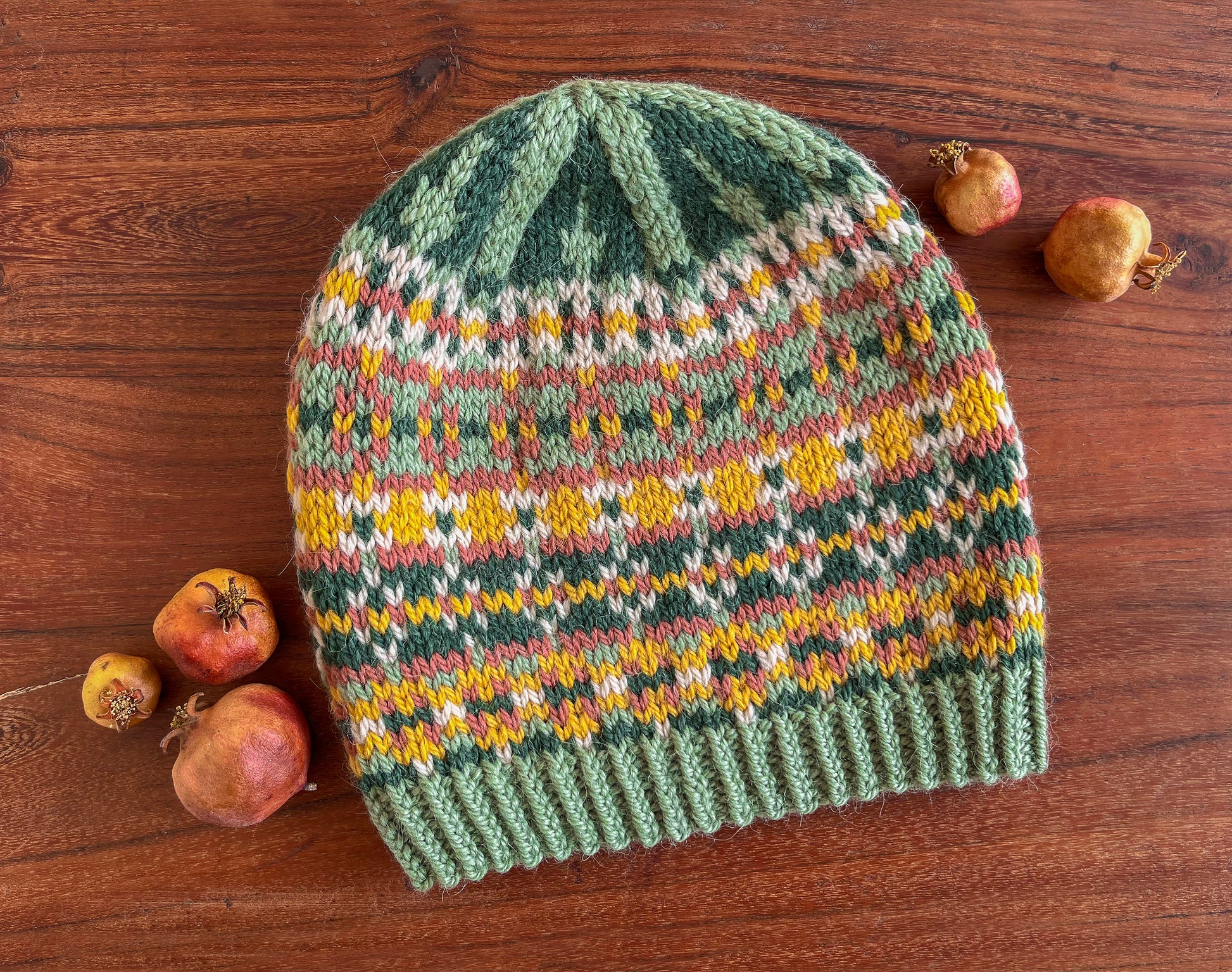 A hand-knitted hat with a Fair Isle pattern in dark green, sage green, yellow, pink, and beige, displayed on a wooden surface with small dried pomegranates around it.