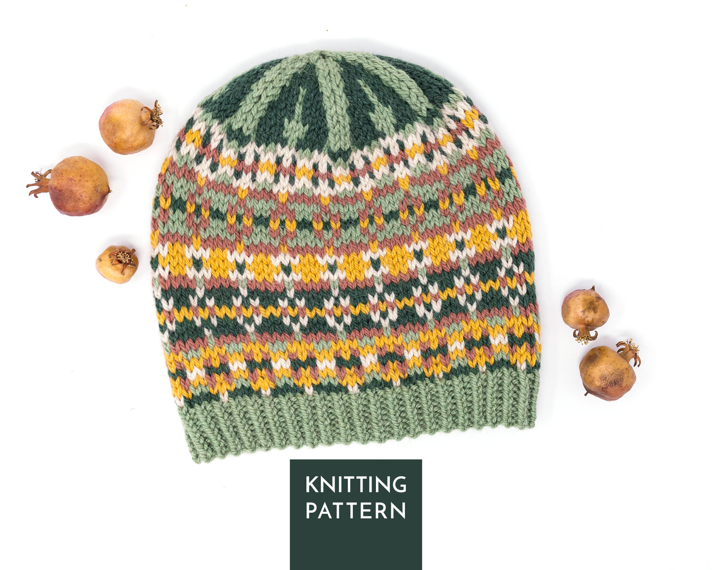 A knitted beanie hat with a Fair Isle style pattern in sage green, dark green, yellow, orange, and beige, styled with dried pomegranates on a white background. A dark green label at the bottom reads 'KNITTING PATTERN'.