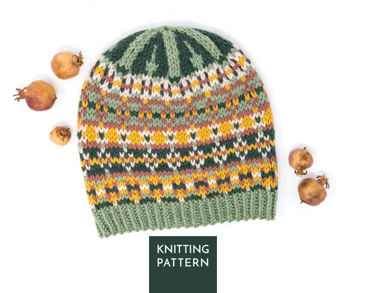 A knitted beanie hat with a Fair Isle style pattern in sage green, dark green, yellow, orange, and beige, styled with dried pomegranates on a white background. A dark green label at the bottom reads 'KNITTING PATTERN'.