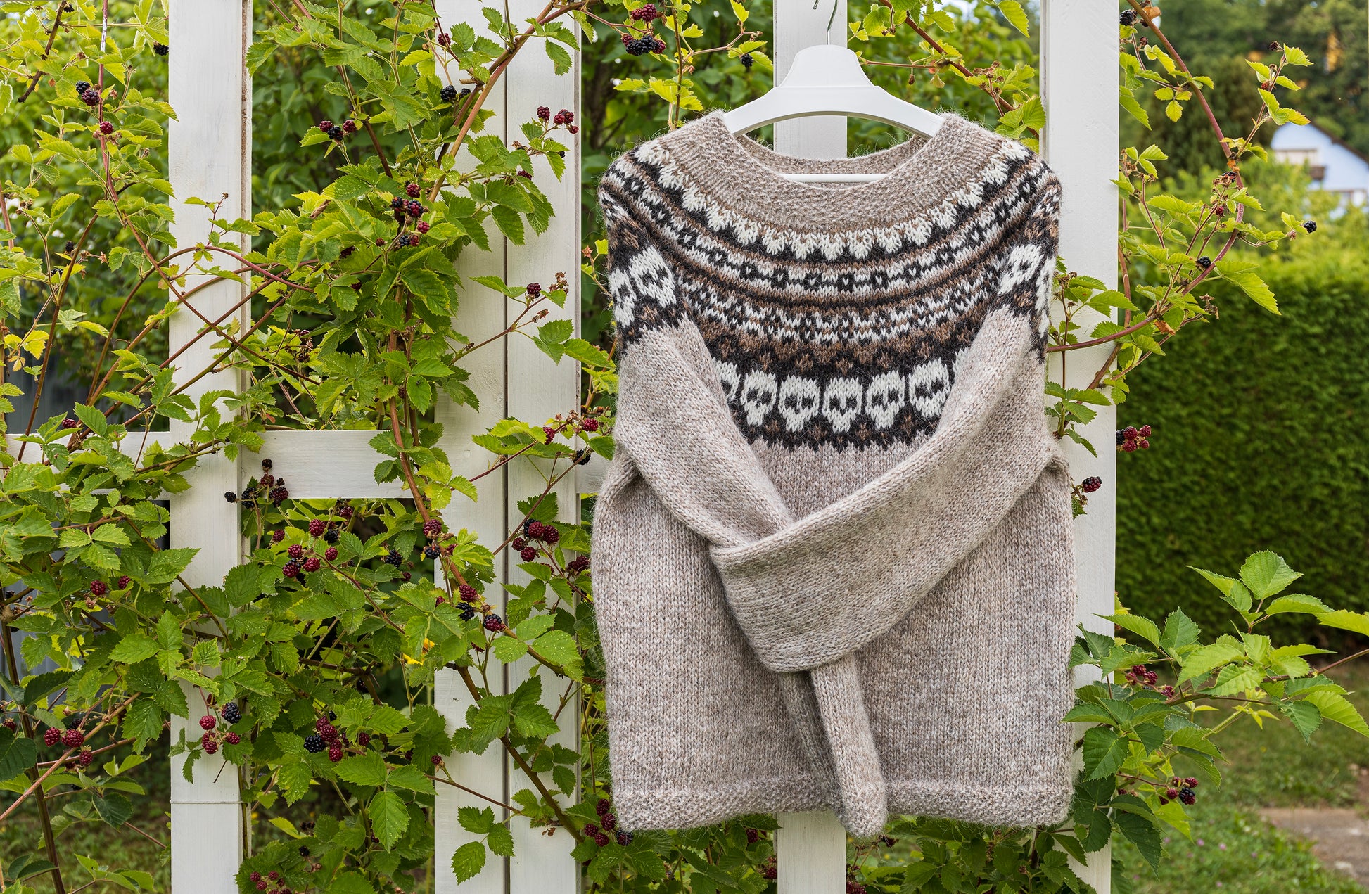 had-knitted lopapeysa sweater with a skull colorwork yoke on a hanger against a white picket fence and greenery background