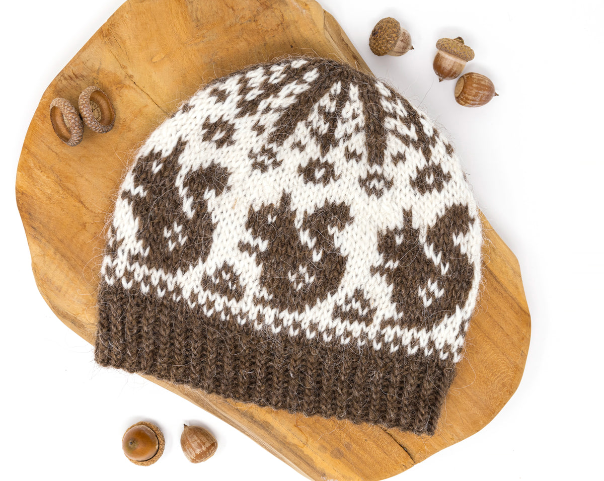 A knitted beanie hat with a brown and off-white Fair Isle squirrels motif pattern, displayed on a rustic wooden slice with acorns