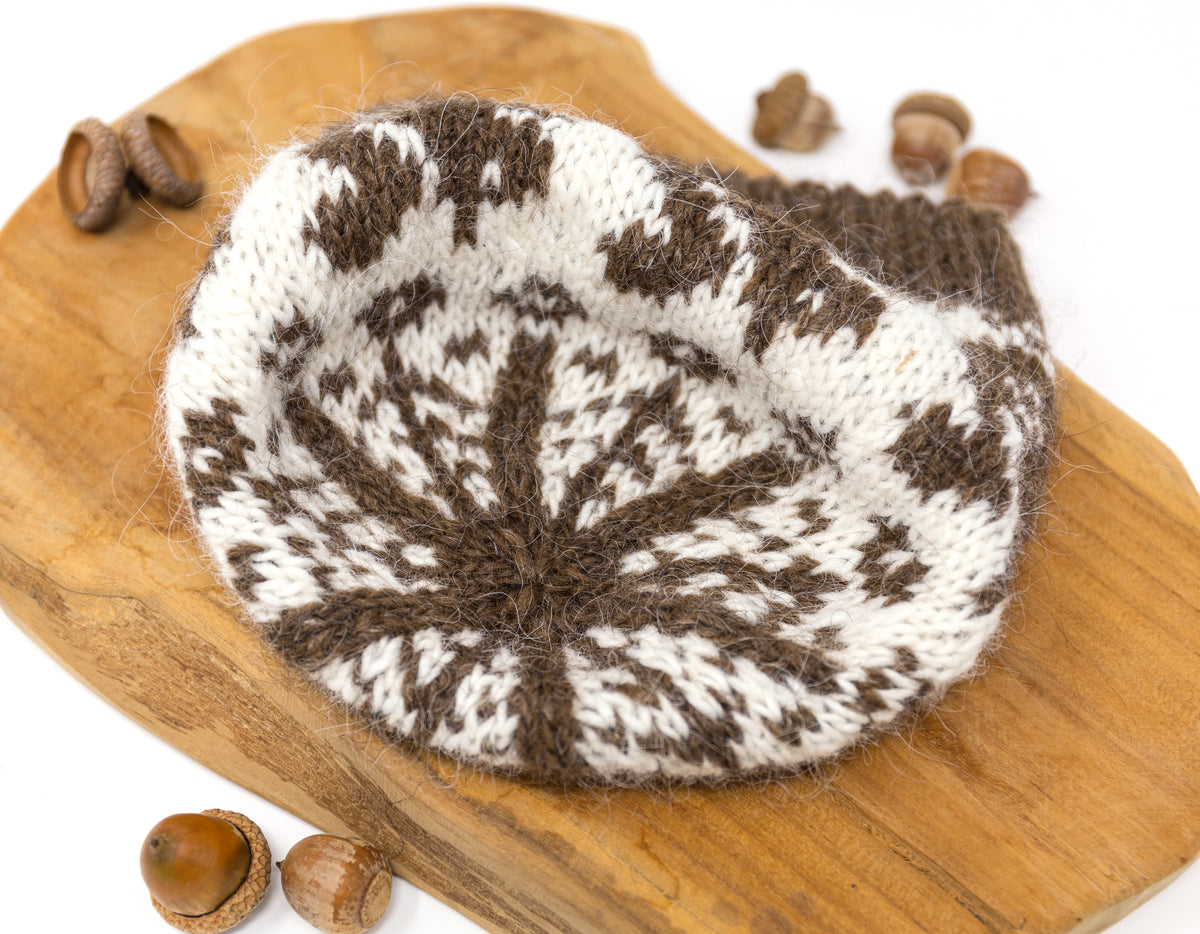 The close up for a corwn of a knitted beanie hat with a brown and off-white Fair Isle squirrels pattern, displayed on a white background with acorns,