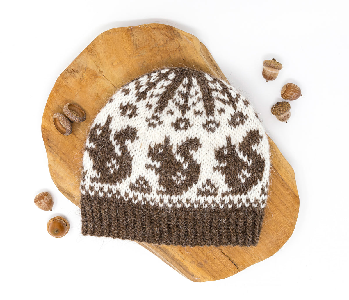 A knitted beanie hat with a brown and off-white Fair Isle squirrels pattern, displayed on a wooden board on a white background with acorns,