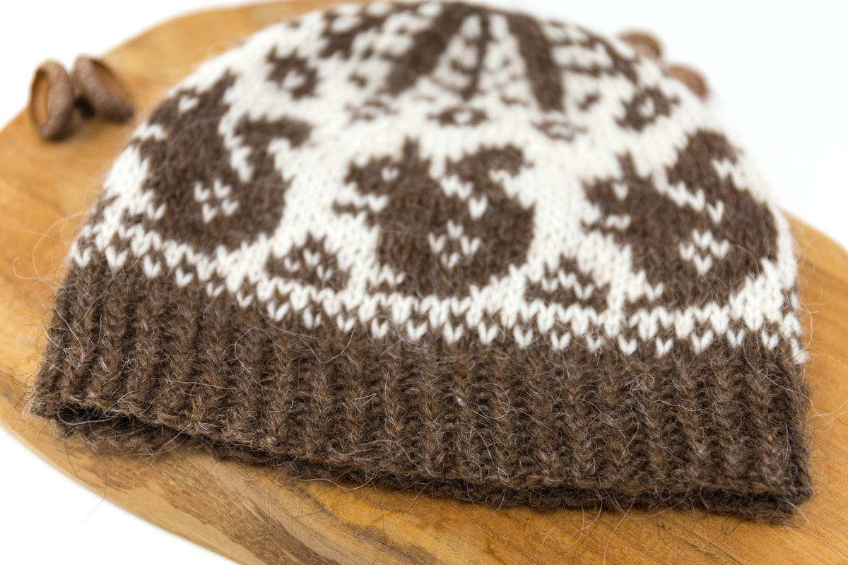 A brown and off white knit beanie hat with a Fair Isle pattern, resting on a wooden board with acorns.