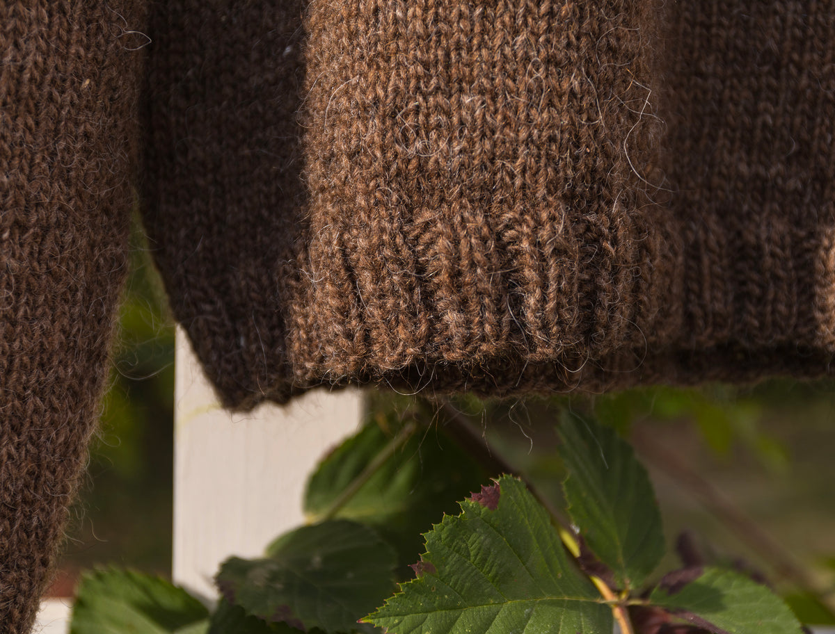 Close-up of a brown knit sweater with a ribbed hem, hanging outdoors with green leaves and a white fence blurred in the background.