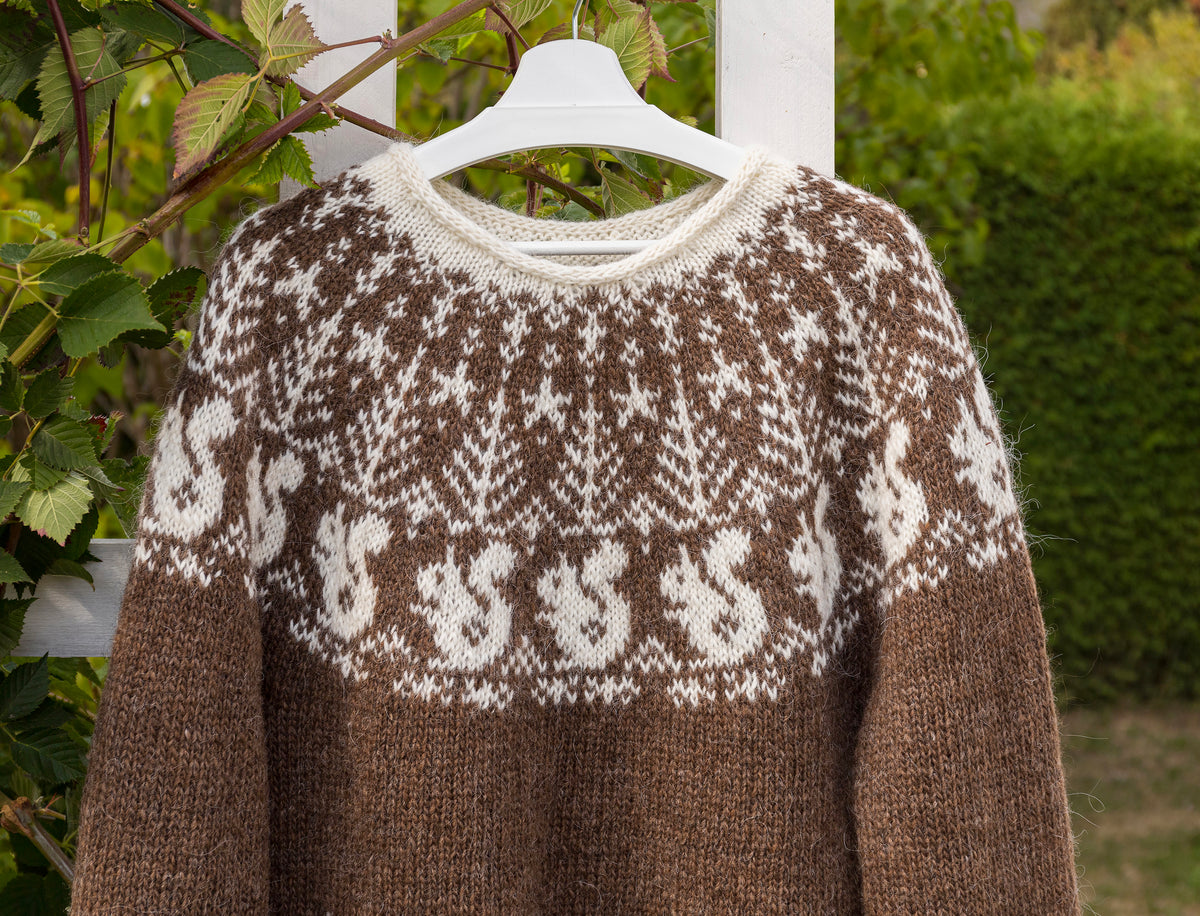 A brown and cream knit sweater with a Fair Isle squirrel pattern, hanging on a white hanger outdoors against a background of green foliage and a white fence.