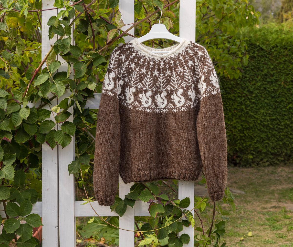 A brown and white hand-knit sweater with a Fair Isle squirrel pattern, hanging on a white hanger against a white wooden fence with green ivy and garden foliage.
