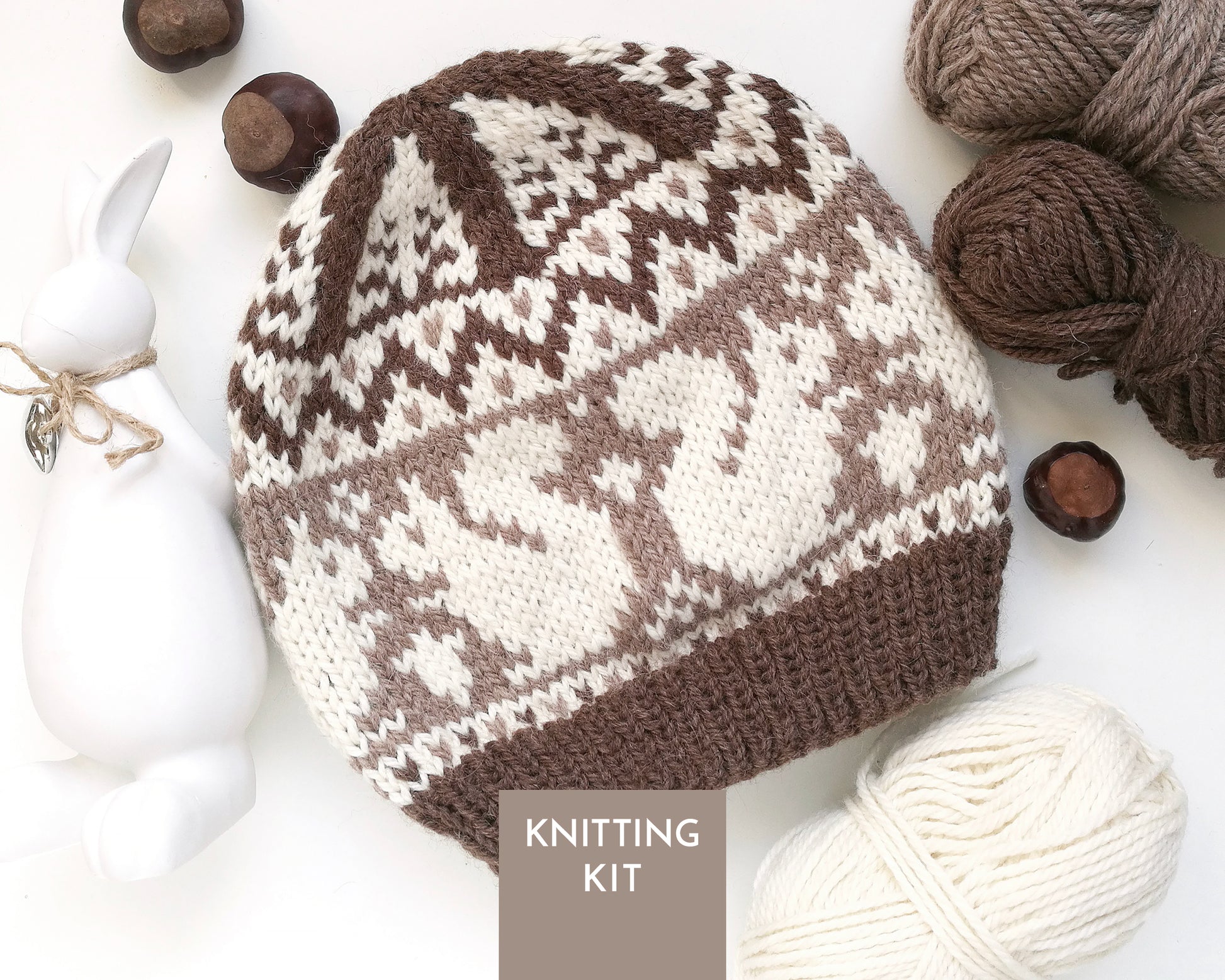 A knitted beanie hat with a brown and white squirrel pattern, styled in a flat lay with yarn balls, acorns, a white ceramic rabbit figurine, and a 'KNITTING KIT' label overlay.