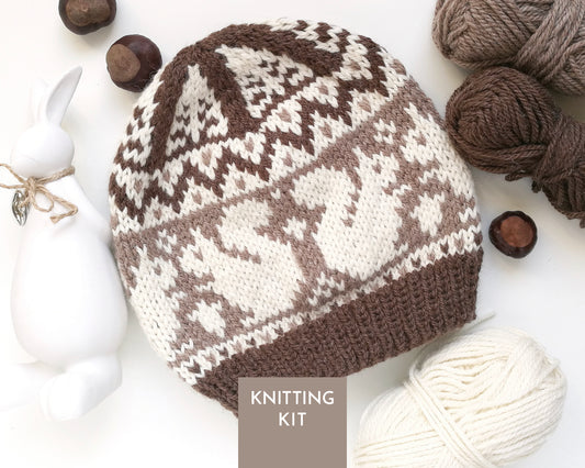 A knitted beanie hat with a brown and white squirrel pattern, styled in a flat lay with yarn balls, acorns, a white ceramic rabbit figurine, and a 'KNITTING KIT' label overlay.