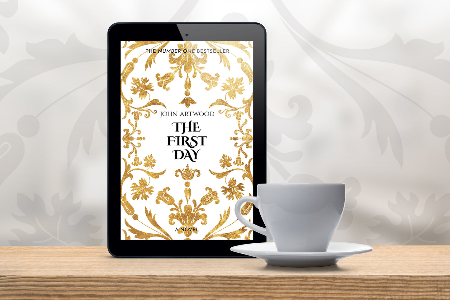 A tablet with a book cover featuring gold baroque ornament, accompanied with a white cup on a wooden table.