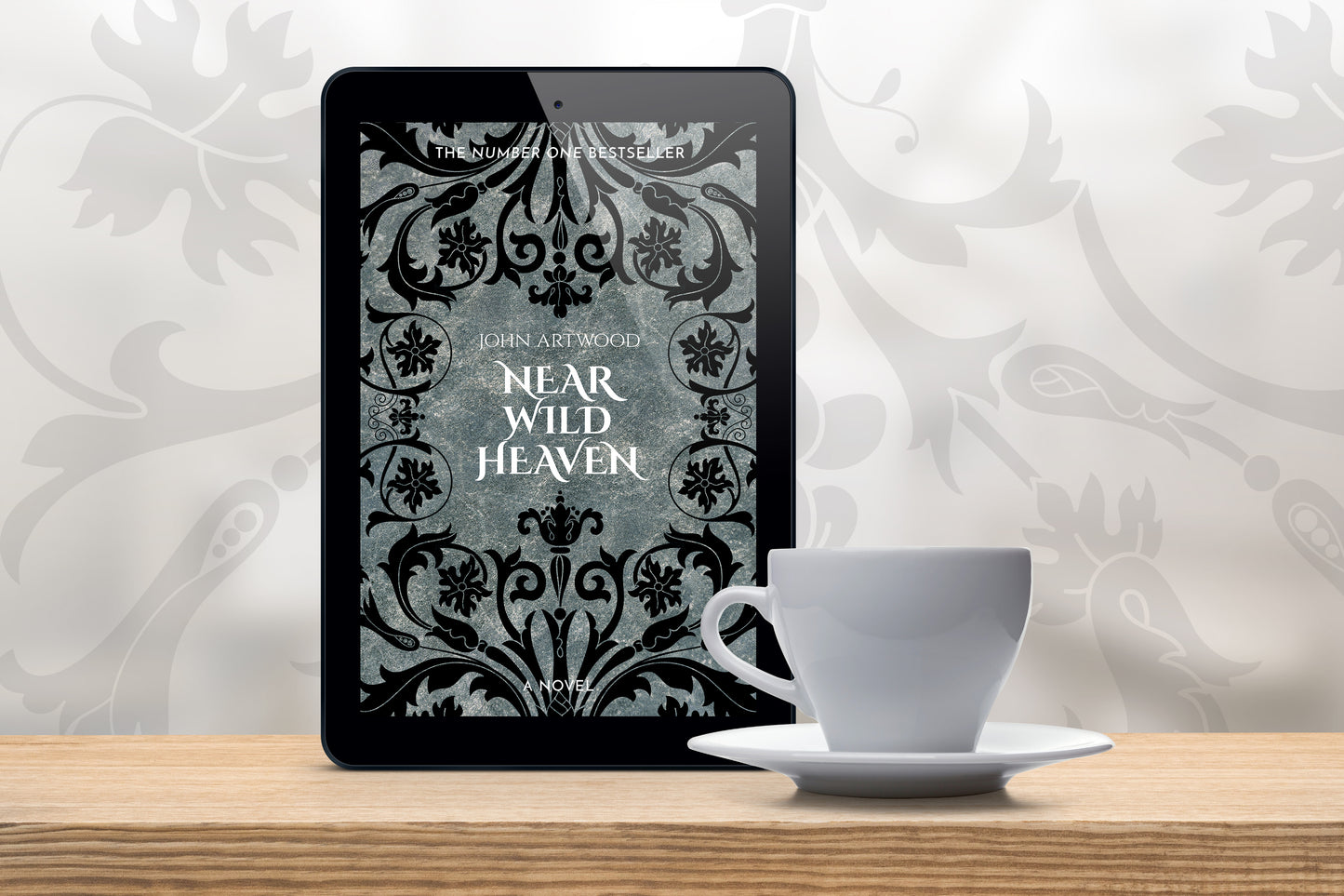 A tablet with a book cover featuring silver and black baroque ornament, accompanied with a white cup on a wooden table.