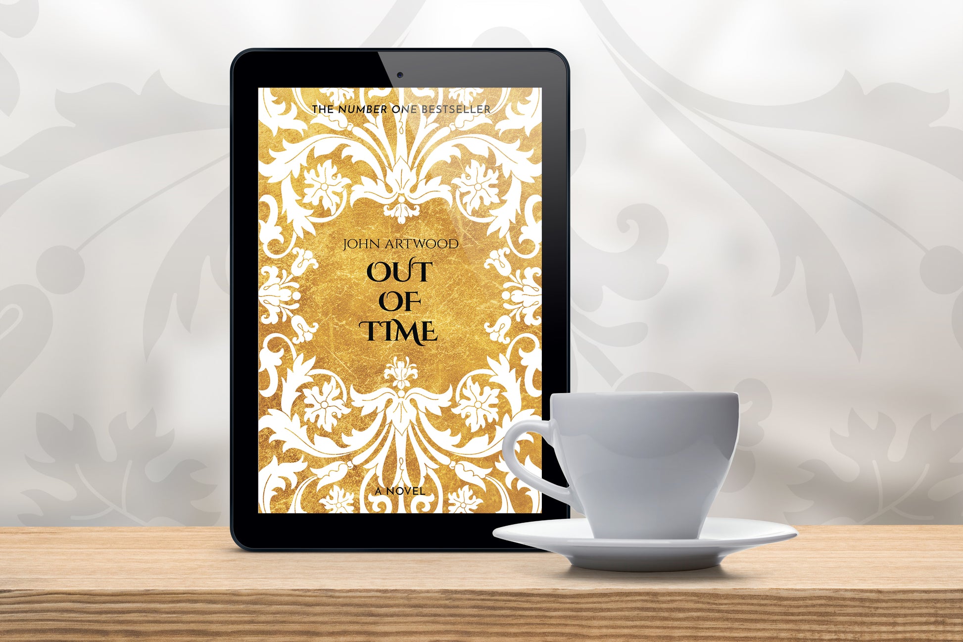 A tablet with a book cover featuring gold and white baroque ornament, accompanied with a white cup on a wooden table.