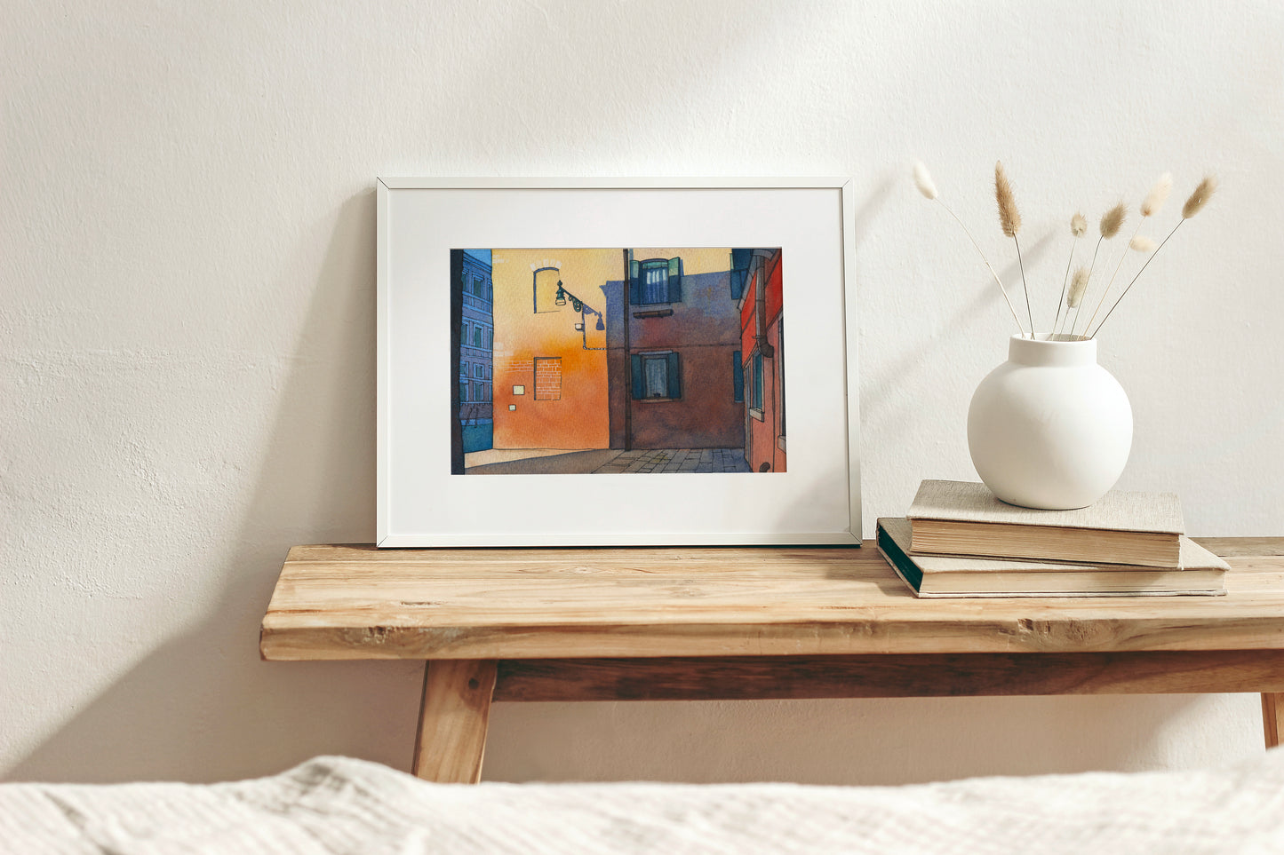 Framed Venice canal street artwork on a wooden shelf with a vase and books against a white wall.