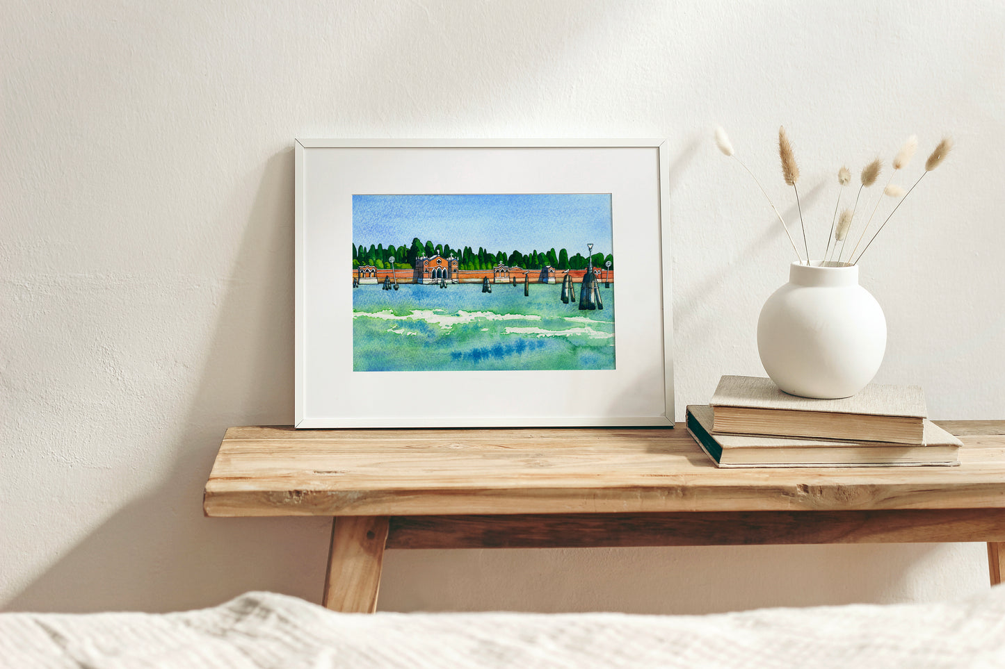 Framed watercolor and ink artwork os San Michele island in Venice on a wooden shelf with a vase and books in the background
