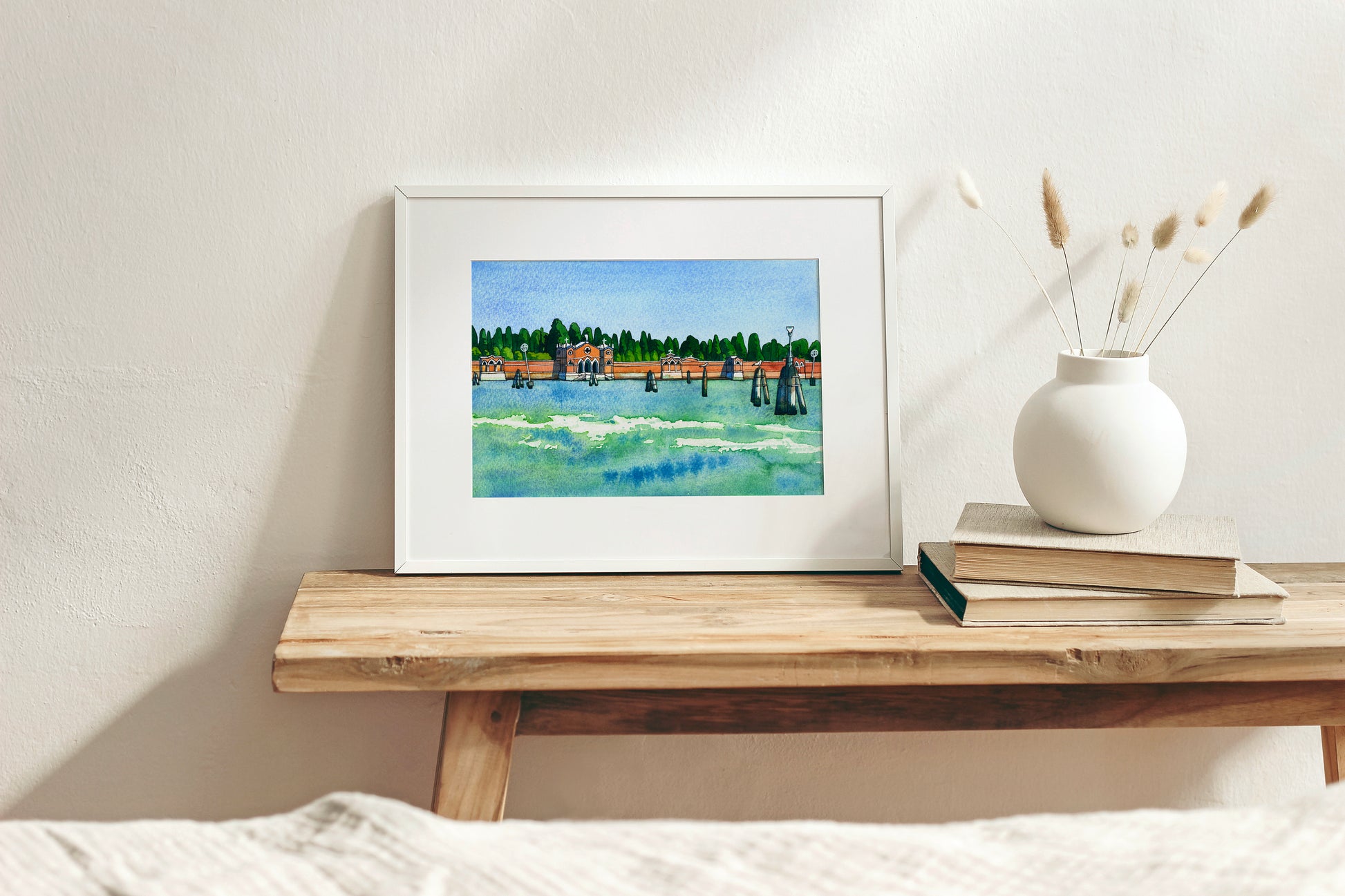 Framed watercolor and ink artwork os San Michele island in Venice on a wooden shelf with a vase and books in the background
