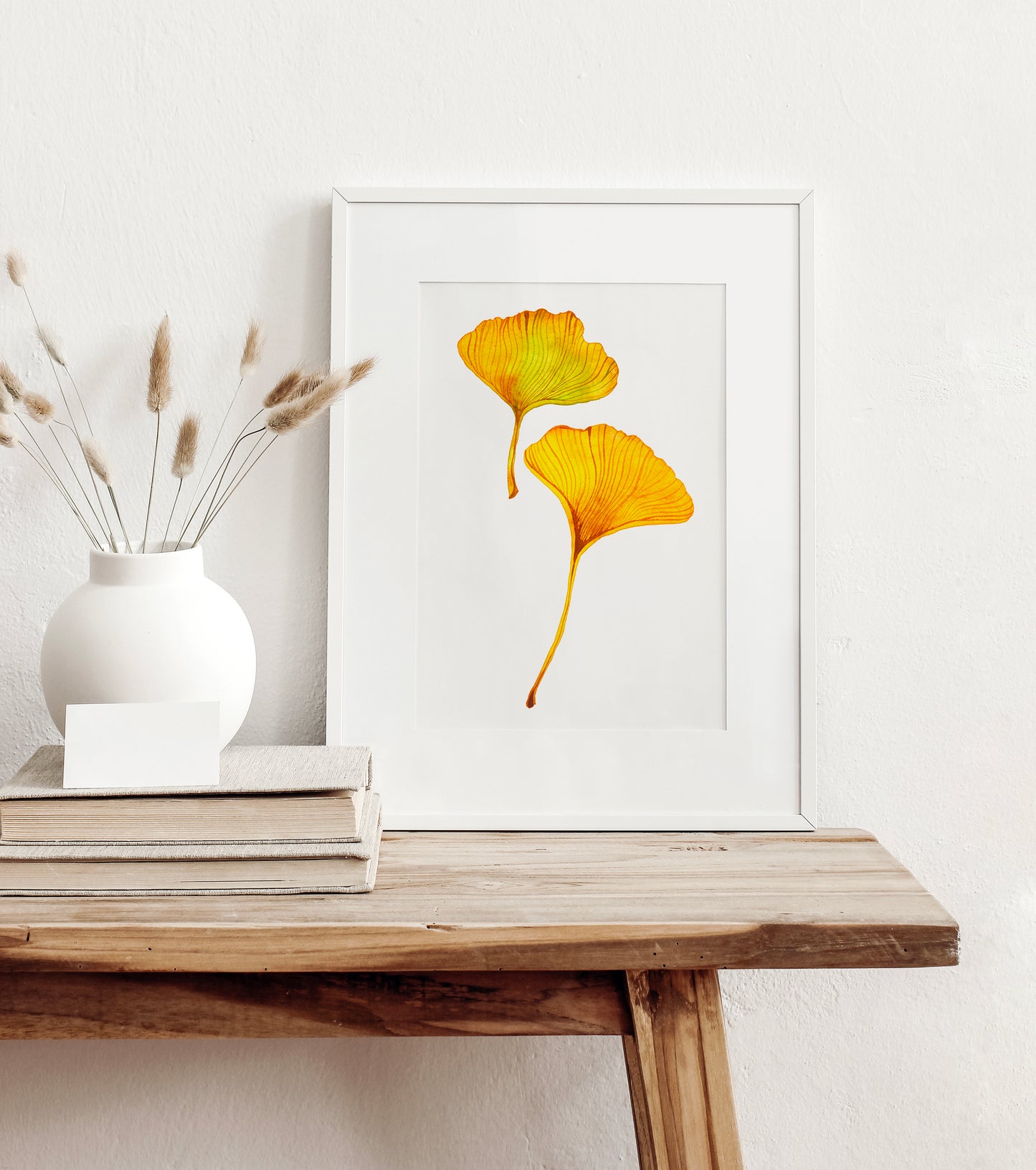 A white-framed botanical watercolor art print featuring two yellow ginkgo leaves on a white background, styled on a rustic wooden table next to a white vase with dried grass stems and stacked books.